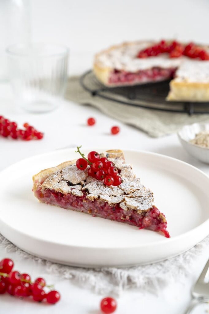 Omas schneller Johannisbeerkuchen – fruchtig und saftig Ein Stück Johannisbeerkuchen und frische rote Johannisbeeren als Dekoration auf einem weißen Teller