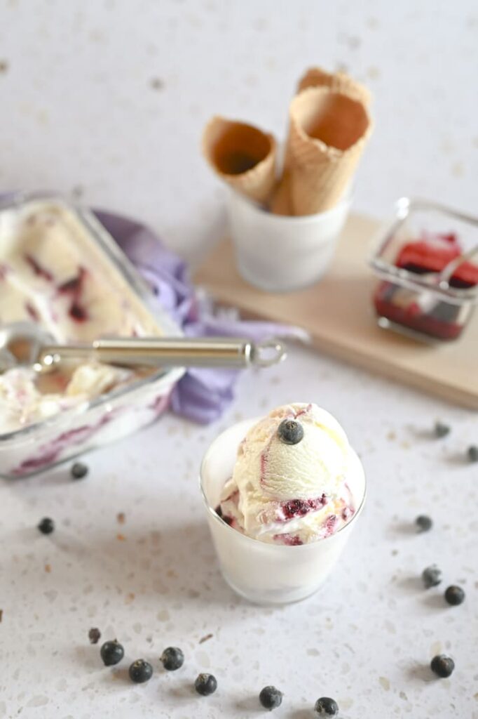 Frozen Joghurt mit Beeren und Johannisbeer-Swirl – cremig und erfrischend Glas mit Frozen Joghurt und Johannisbeer-Swirl, garniert mit einer Beere, im Hintergrund eine Eisschale, Waffeln und Fruchtsoße