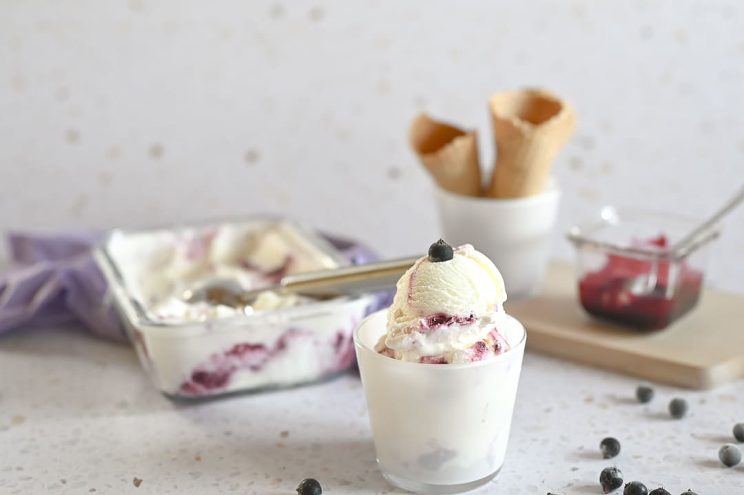 Frozen Joghurt mit Johannisbeer-Swirl in einem Glas, im Hintergrund eine Auflaufform mit Eis und Eiswaffeln