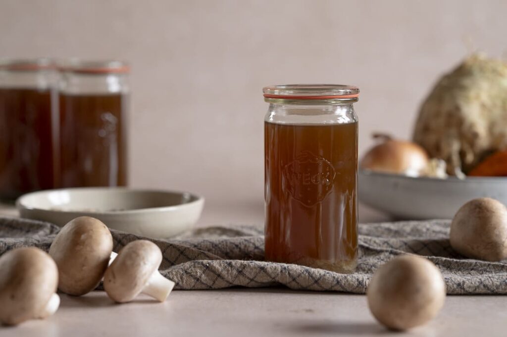 Glas mit selbstgemachter Pilzbrühe auf Küchentuch – frische Champignons im Vordergrund, Herbstgemüse im Hintergrund.