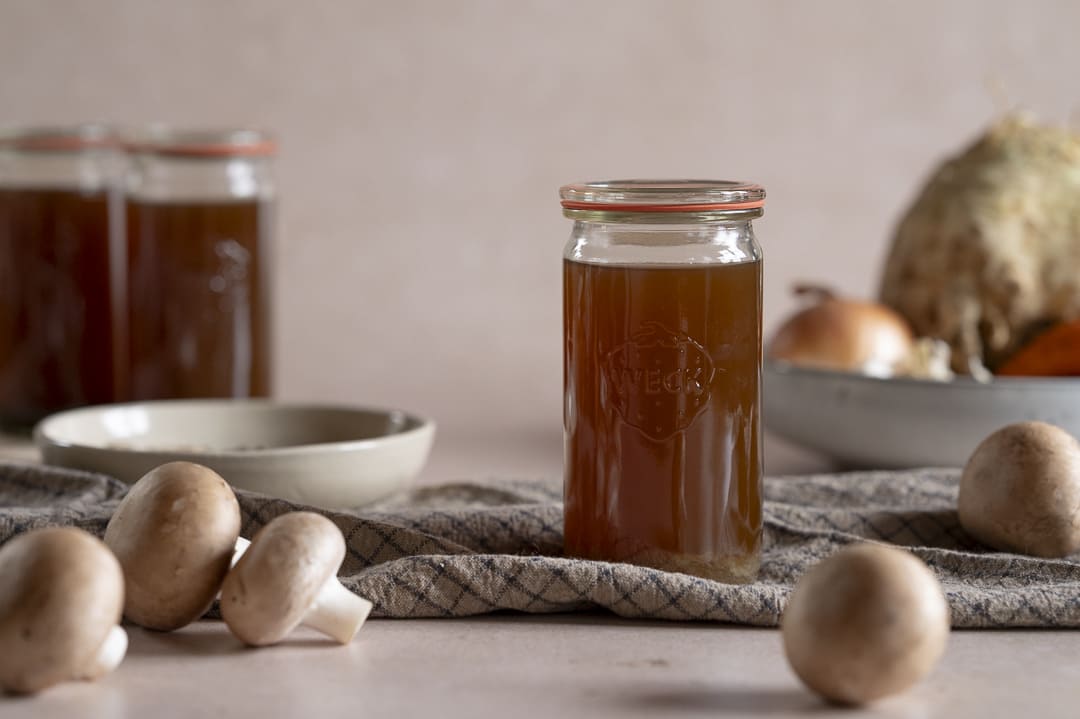 Glas mit selbstgemachter Pilzbrühe auf Küchentuch – frische Champignons im Vordergrund, Herbstgemüse im Hintergrund.